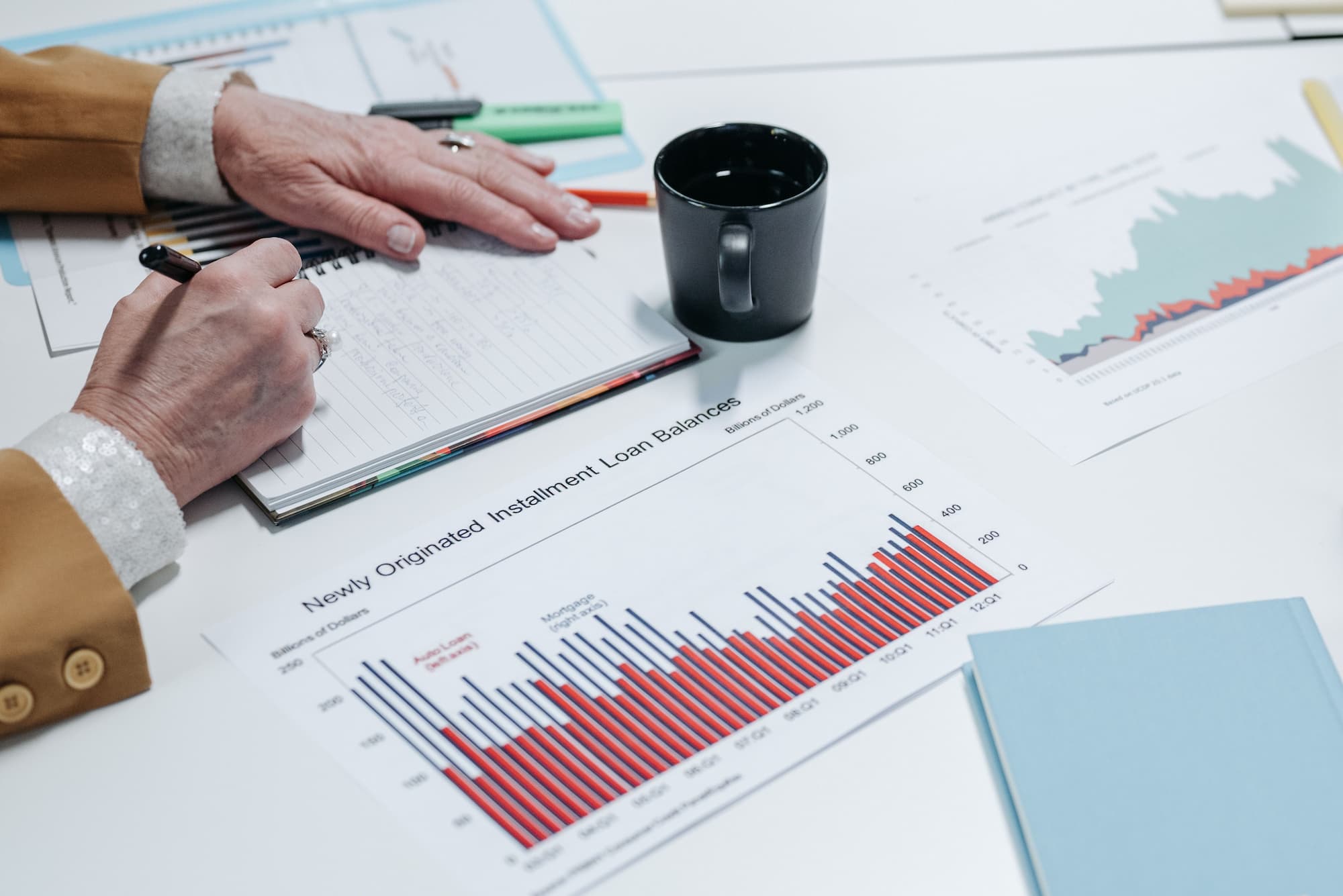 Close-up of a hand writing notes beside financial charts and reports spread across a desk.